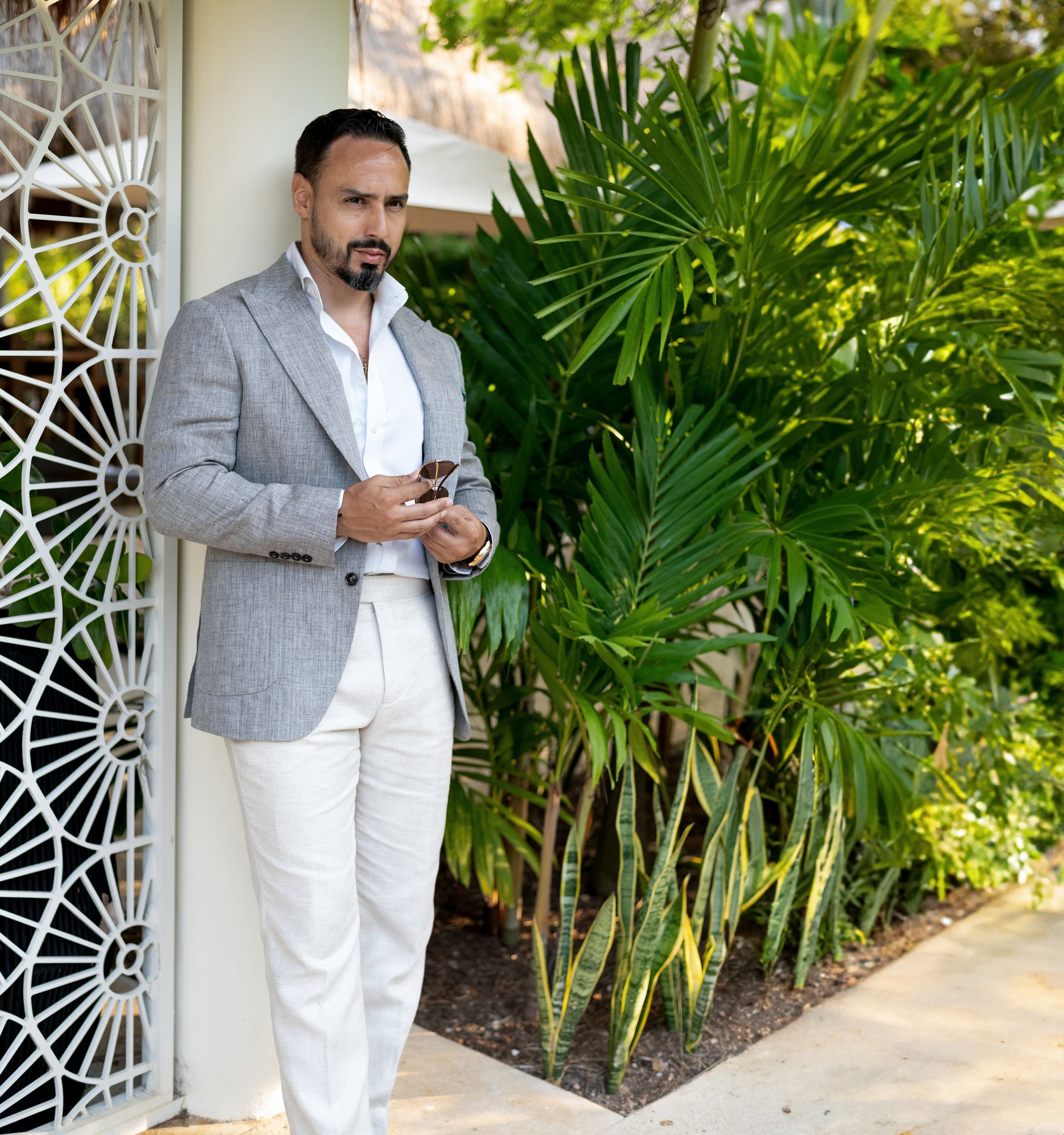 César Renuan in a grey linen jacket and white trousers beside a lattice screen and palms